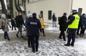 Policjantka stojąca tyłem rozdaje przechodniom odblaski i ulotki informacyjne. W tle widać pieszych oraz ścianę budynku urzędu Miasta i gminy w Olkuszu.