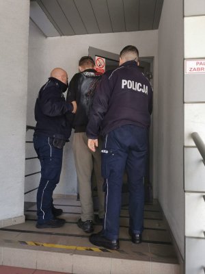Zatrzymany 27-letni mężczyzna doprowadzany do budynku Komendy Powiatowej Policji w Olkuszu. Po obu stronach umundurowani policjanci. Osoby stoją tyłem.