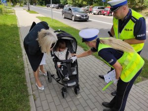 Policjantka i policjant wręczają ulotki kobiecie. Policjantka nachyla się nad wózkiem dziecięcym i przekazuje odblask dziewczynce.