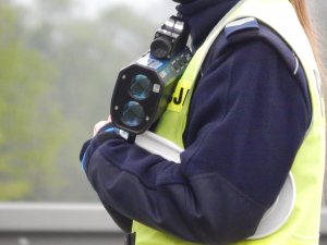 Policjantka ruchu drogowego w kamizelce odblaskowej trzymająca przy sobie urządzenie do pomiaru prędkości kierujących pojazdami tak zwany prędkościomierz.