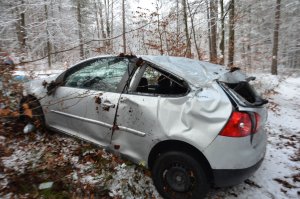 Srebrny volkswagen golf po wypadku. Zniszczona karoseria, wybite szyby w pojeździe. Samochód znajduje się w lesie wokół samochodu drzewa.