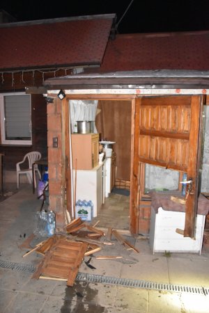 Uszkodzone drewniane drzwi znajdujące się przy  wejściu do budynku (ganku). Porozrzucane elementy desek  znajdują się na kostce brukowej. Za drzwiami widać meble kuchenne i inny sprzęt.