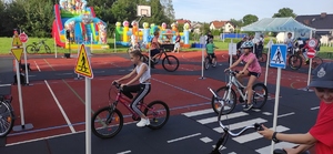 Dzieci jeżdżą na rowerach po miasteczku ruchu drogowego. W  tle kolorowe dmuchańce. Pogoda letnia, słoneczna.