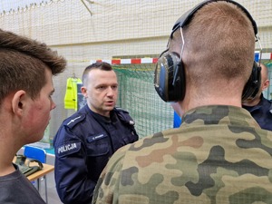 Policjant z dwoma uczniami klasy o profilu mundurowych. Mężczyźni stoją tyłem. Jeden z nich ma słuchawki na uszach.