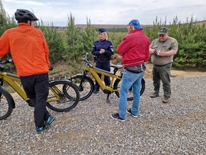 Policjantka i funkcjonariusz straży leśnej podczas legitymowania dwóch rowerzystów. W tle pejzaż, lasy i piaski