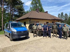 Policjanci i funkcjonariusze straży leśnej przy pojazdach służbowych. w tle budynek oraz krajobraz leśny