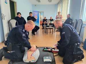 Policjanci ćwiczą pierwszą pomoc  na fantomie przy użyciu defibrylatora. W tle inni funkcjonariusz