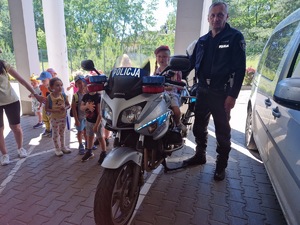 Dziecko na policyjnym motocyklu obok policjant. w oddali grupa przedszkolaków