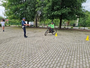 Dziecko na boisku szkolnym na rowerze robi ósemkę obok policjant ruchu drogowego (biała czapka)  dr