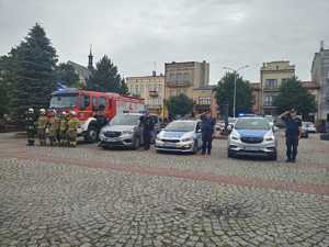 Funkcjonariusze Straży Pożarnej, Straży Miejskiej oraz policjanci przy pojazdach służbowych oddają hołd poległemu żołnierzowi. W tle rynek i zabudowania.