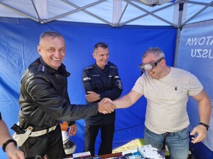 Mężczyzna w alkogoglach podaje rękę policjantowi. Obok stoi drugi policjant. Zdjęcie przy stanowisku profilaktyczny. Na stoliku broszury, ulotki gadżety