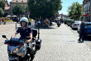 Policjanci ruchu drogowego na motocyklach zabezpieczają przemarsz. W tle ludzie, olkuski rynek i zabudowania. Pogoda letnia słoneczna.  oraz