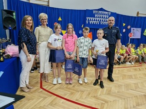 Dyrektor, zastępca Dyrektora szkoły oraz I Zastępca Komendanta Powiatowego Policji w Olkuszu wraz z laureatami konkursu. W tle dekoracji , znaki drogowe oraz młodzież w odblaskowych kamizelkach.