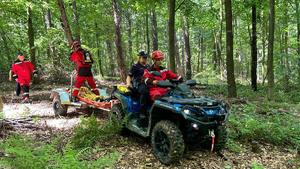 Strażak oraz policjantka na quadzie jada przez las.Quad ciągnie przyczepę na której stoi strażak.Za quadem idzie kolejny strażak w czerwonym umundurowaniu . Strażacy na quadzie w kaskach ochronnych na głowach.