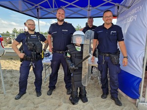 Policjanci przy chłopcu. Chłopiec ma założony sprzęt przeciw uderzeniowy, stoi w kasku w rękach trzyma tarczę i pałkę.