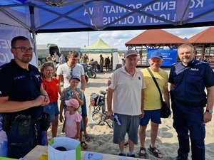 Grupa osób gromadzi się przy stanowisku policyjnym pod namiotem przy policjantach. W tle namioty i uczestnicy imprezy