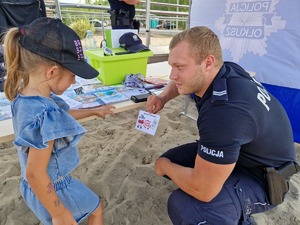 Policjant kuca przy dziewczynce pokazuje jej numer alarmowy. W tle stanowisko profilaktyczne, broszury, ulotki i gadżety.