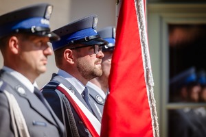 Poczet sztandarowy. Trzech policjantów stoi w rzędzie, w środku policjant trzyma sztandar.