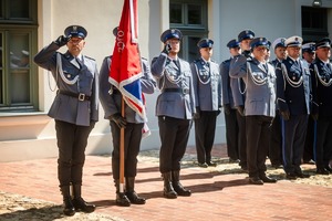 Poczet sztandarowy oraz policjanci stoją w równym rzędzie oddają honory. W tle fragment budynku