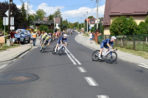 Peleton rowerzystów wyścigu Tour de Pologne. Na tle zabudowań. Przy drodze gromadzą się gapie.