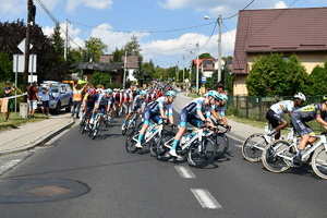 Peleton rowerzystów podczas Tour de Pologne. W tle zabudowania. Przy ulicy na chodniku gromadzą się gapie. Po prawej stronie radiowóz policyjny