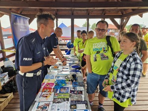 Stanowisko profilaktyczne policji. Dwóch policjantów, przy stolikach. Na przeciwko uczestnicy imprezy, dzieci z rodzinami. Na stolikach ulotki, plakaty broszury, kolorowe malowanki, .