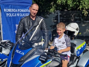 Policjant ruchu drogowego stoi przy chłopcu, który siedzi na policyjnym motocyklu. Zdjęcie przy stanowisku profilaktycznym Policji.