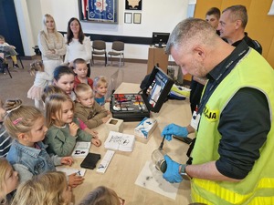 Technik kryminalistyki pokazuje dzieciom jak zebrać odciski linii papilarnych. Grupa dzieci gromadzi się przy policjancie