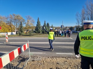 Policjantka ruchu drogowego w odblaskowej kamizelce kieruje ręcznie ruchem na drodze. W tle widać pieszych.