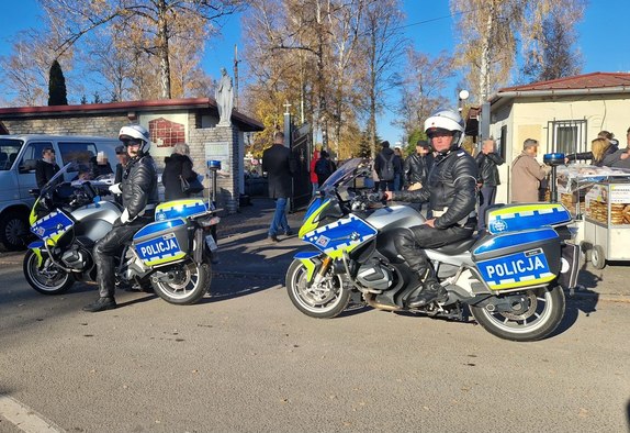 Dwaj policjanci ruchu drogowego na motocyklach przy cmentarzu. w tle ludzie odwiedzający cmentarz