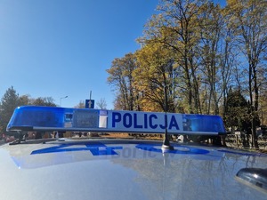 Fragment radiowozu. Na dachu światła policyjne z napisem POLICJA. W tle pejzaż i drzewa. Pogoda jesienna. Na drzewach żółte i pomarańczowe liście.