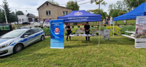 Policjanci na stanowisku profilaktycznym Komendy Powiatowej Policji w Olkuszu, obok radiowóz policyjny.