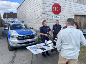 Policjanci z lokalna społecznością przy stanowisku profilaktycznym. W tle radiowóz policyjny.