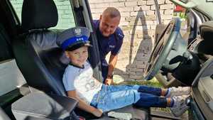 Dziecko w radiowozie policyjnym w czapce policjanta. W tle policjant.