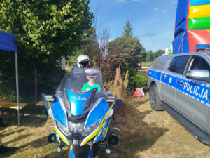 Dziecko w czapce policjanta Ruchu Drogowego na motocyklu. Po prawej stronie radiowóz policyjny.