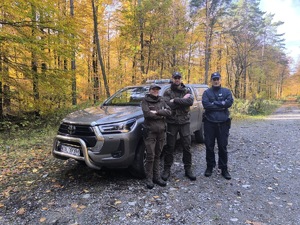Policjant i funkcjonariusze Straży Leśnej, w tle pojazd służbowy Straży Leśnej.