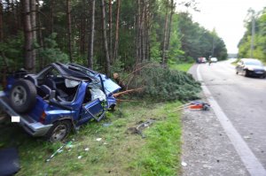 Niebieski samochód suzuki vitara po zderzeniu z drzewem. Przód samochodu rozbity, otwarta klapa, w tle złamane drzewo i las. Po prawej stronie droga asfaltowa.