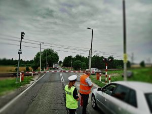 Policjantka i pracownik PKP wręczają kierowcy ulotkę informując o zagrożeniach. Samochód znajduje się przed przejazdem kolejowym.