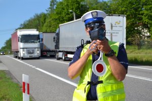 Policjant ruchu drogowego  w mundurze i odblaskowej kamizelce dokonuje pomiaru prędkości kierowców urządzeniem to jest prędkościomierzem. W tle widać drogę i jadące samochody.