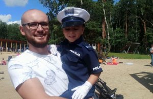 Mężczyzna trzyma na rękach dziecko ubrane w mały policyjny strój. Dziecko ma na głowie białą czapkę policjanta ruchu drogowego.