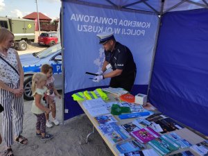 Stanowisko profilaktyczne pod namiotem policji z ulotkami i informacjami przy którym policjant ruchu drogowego wręcza dzieciom odblaski.