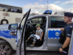 W radiowozie siedzi dziecko. Trzyma w ręce malowankę. Obok stoi policjant.