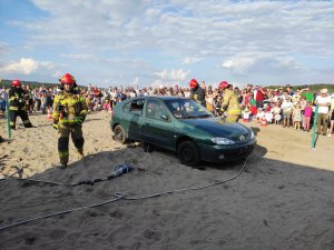 Strażacy rozcinają drzwi samochodu za pomocą specjalistycznego sprzętu. Dookoła gromadzą się uczestnicy imprezy.