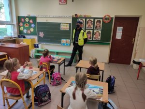 Policjant ruchu drogowego na prelekcji w Sali w szkole podstawowej rozmawia z uczniami. Dzieci siedzą pojedynczo, w odstępach w szkolnych ławkach.