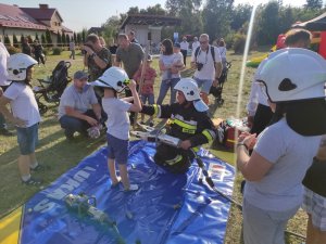 Chłopiec w kasku strażackim stoi przy strażaku, który trzyma w ręce specjalistyczne narzędzie, wykorzystywane podczas wypadków drogowych.  W tle gromadzą się zainteresowani widzowie.