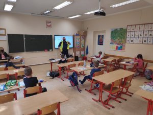 Policjant w kamizelce odblaskowej prowadzi zajęcia w klasie. Dzieci siedzą w odstępach w szkolnych ławkach. Zdjęcie wykonane w klasie.