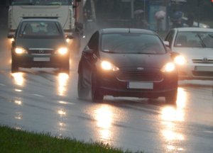 Perspektywa jezdni o trzech pasach ruchu w jednym kierunku, po której jadą (na wszystkich tych pasach) samochody z włączonymi światłami mijania. Przejrzystość powietrza obniżona - pada deszcz. jezdnia jest mokra i śliska - odbija światła reflektorów.