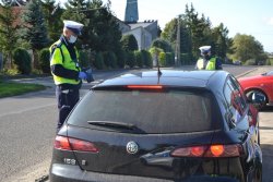 Policjant  ruchu drogowego w białej czapce podczas kontroli drogowej. Obok czarny samochód osobowy. W tle droga i drzewa.