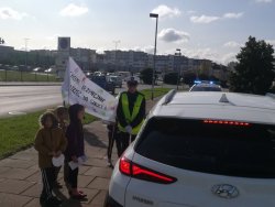 Biały samochód osobowy,  obok policjant ruchu drogowego z dziećmi, które trzymają w ręce transparent i ulotki.  W tle radiowóz.