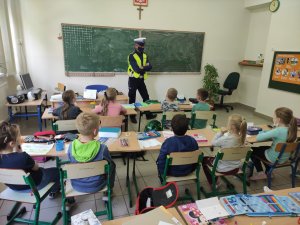 Policjant ruchu drogowego w białej czapce i kamizelce odblaskowej rozmawia z dziećmi zajęcia prowadzone są w klasie lekcyjnej. Dzieci siedzą w szkolnych ławkach.